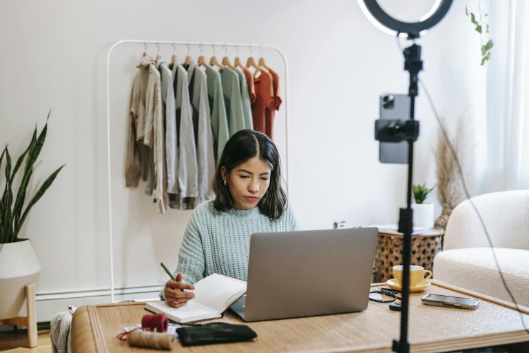 Focused young woman working on laptop in home office, surrounded by clothing rack and camera setup.