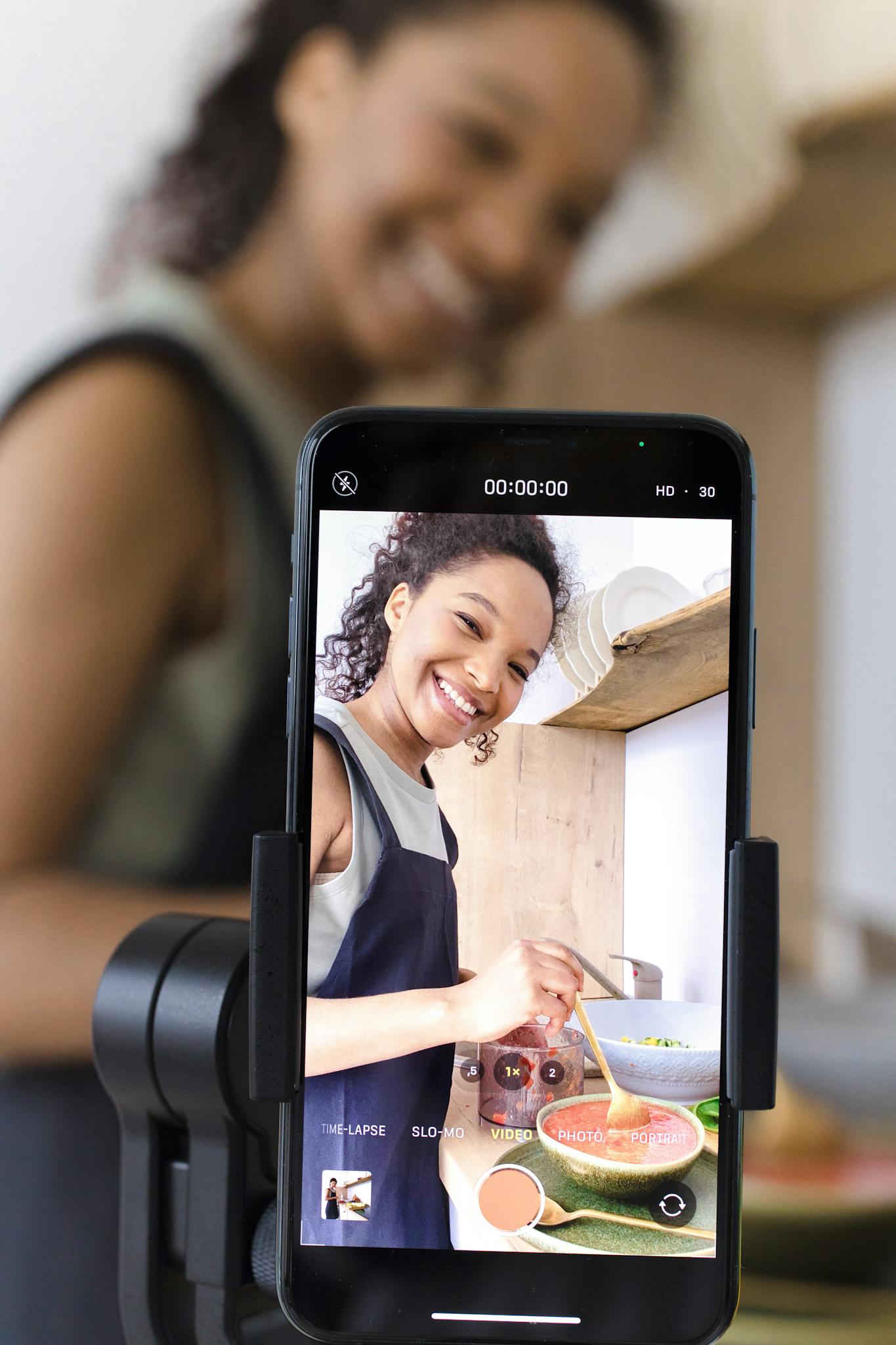 Smiling woman filming cooking video on smartphone in kitchen.